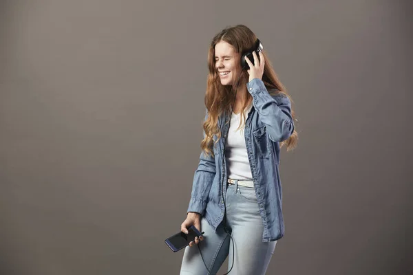 Sonriente chica de cabello castaño vestida con una camiseta blanca, jeans y camisa vaquera sostiene el teléfono en su mano y escucha algo por los auriculares sobre un fondo gris — Foto de Stock