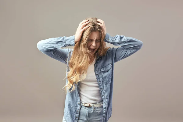 Triest bruinharige meisje gekleed in een wit t-shirt en jean houdt haar handen op haar hoofd en schreeuwt op een grijze achtergrond — Stockfoto