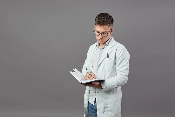 Tipo inteligente en gafas vestidas con una camisa blanca y abrigo blanco hace notas en un cuaderno sobre un fondo gris — Foto de Stock