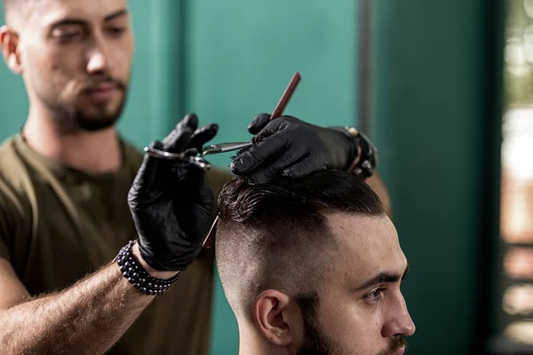 Barber in black gloves cuts with scissors hair of stylish man at a barbershop