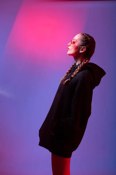 Portrét, fashion Girl v černý svetr s kapucí a sluneční brýle kulatý tvar v neonové světlo v ateliéru — Stock fotografie