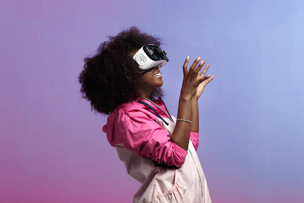 Modern curly brown-haired girl dressed in the pink sports jacket uses the virtual reality glasses in the studio on neon background