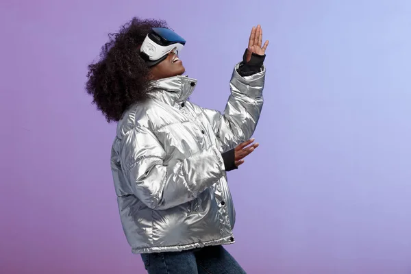 Mod chica de pelo castaño rizado vestido con una chaqueta de color plateado utiliza las gafas de realidad virtual en el estudio sobre fondo de neón — Foto de Stock