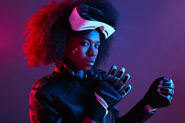 Stylový kudrnaté černovlasá dívka na sobě černou koženou bundu a rukavice pózy s virtuální realita brýle na hlavu v temné studio s neon light — Stock fotografie