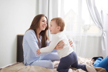Genç güzel anne ve küçük oğlu pijama giymiş hafif rahat yatak odasında yatağın üstüne iyi eğlenceler
