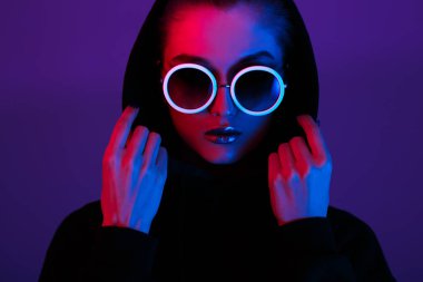 Siyah kazaklı bir kukuleta ile ve güneş gözlüğü kırmızı ve mavi neon ışık stüdyo yuvarlak genç kız moda portresi