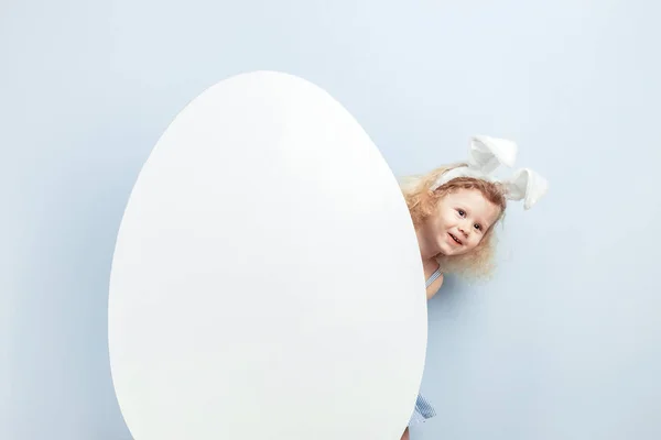 Niña rizada con orejas de conejo en la cabeza escondida detrás de un gran huevo blanco contra una pared azul. Conejo de Pascua — Foto de Stock