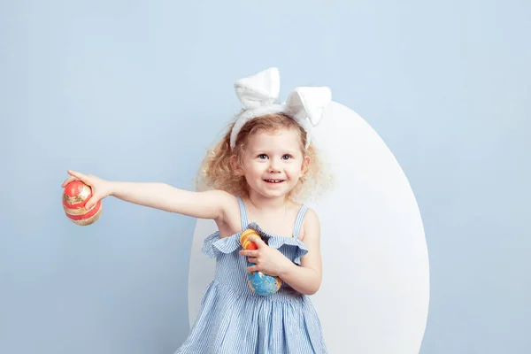 Bájos lány göndör a világoskék ruhát nyuszi füle a fején tartja a kezében egy nagy fehér alapon festett tojás tojás kék falra. Húsvéti nyuszi — Stock Fotó