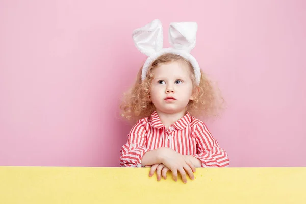 Lustiges kleines, lockiges Mädchen in einem rot-weißen gestreiften Kleid und Hasenohren auf dem Kopf steht hinter der gelben Tafel vor einer rosa Wand — Stockfoto