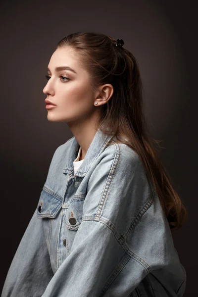 Portrait of beautiful brunette girl with hair tied back dressed in jeans jacket on the dark background in the studio