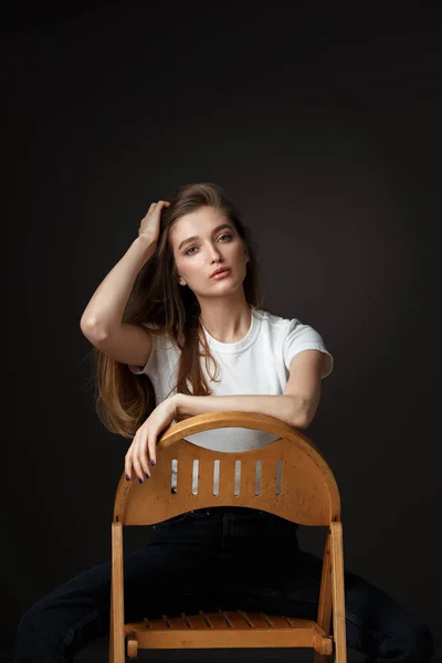 Красива брюнетка дівчина з довгим волоссям, одягнена в білу футболку і джинси, сидить на дерев'яному стільці на чорному фоні в студії — стокове фото
