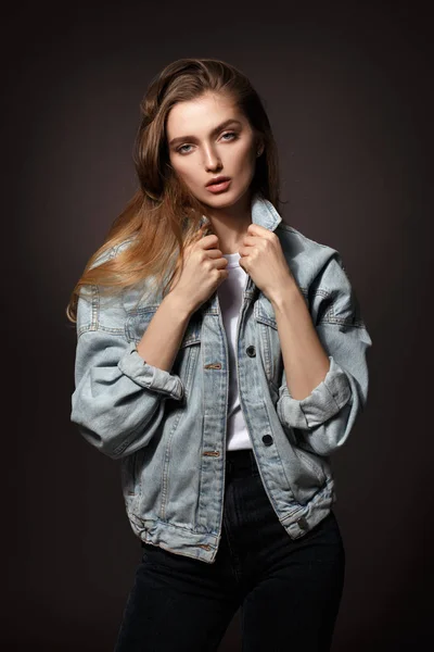 Gorgeous brunette girl with long flowing hair dressed in jeans jacket and jeans poses standing on the dark background in the studio — Stock Photo, Image