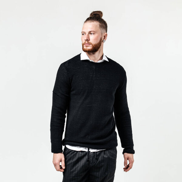 Stylish man with beard and bun hairstyle wearing a black jumper over a white shirt and trousers poses in the studio on the white background
