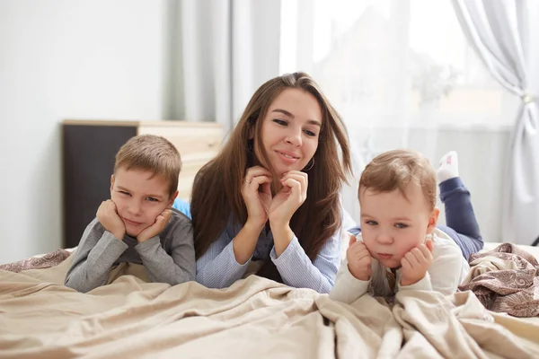 Szczęśliwa młoda matka, ubrana w piżamy niebieskie światło kładzie z jej dwóch synów mały, trzymając ręce pod ich podbródki na łóżku z beżowy kocyk w sypialni z dużym oknem — Zdjęcie stockowe