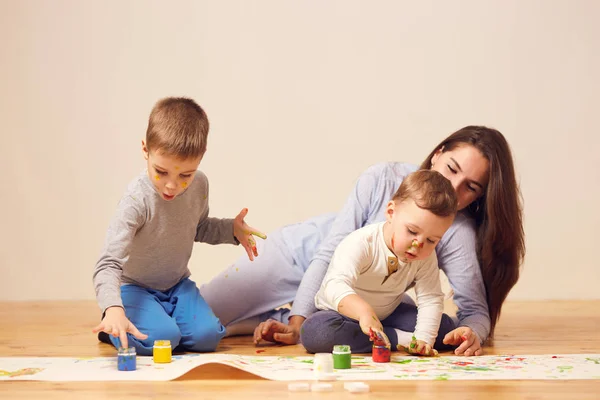 Belle mère heureuse et ses deux petits fils habillés en vêtements de maison sont assis sur le sol en bois dans la chambre et peindre avec les doigts sur le papier blanc — Photo