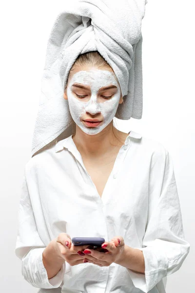 Красивая молодая женщина в белой одежде с белым полотенцем на волосах и косметической маской на лице держит мобильный телефон в руках на белом фоне — стоковое фото