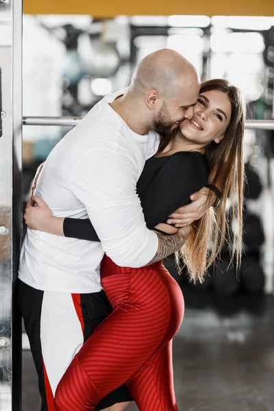 Romántica pareja atlética feliz. Hombre fuerte y esbelta hermosa chica se están abrazando en el moderno gimnasio al lado del equipo deportivo —  Fotos de Stock