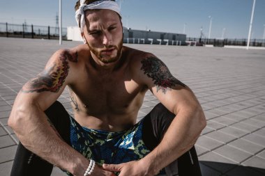 Kafa bandı ve dövmeler ile çıplak bir gövde ile genç atletik erkek siyah tayt giymiş ve mavi şort oturur üzerinde kaldırım döşeme sıcak güneşli bir günde