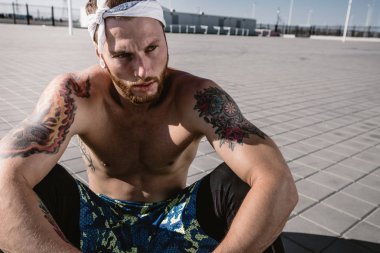 Kafa bandı ve dövmeler ile çıplak bir gövde ile genç atletik erkek siyah tayt giymiş ve mavi şort oturur üzerinde kaldırım döşeme sıcak güneşli bir günde