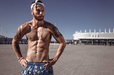 Açık havada egzersiz zeminde eğitimden sonra rahatlatıcı. Yakışıklı gömleksiz sporcu uzakta müzik kentsel poz dinleme earphones ile parlak güneşli bir günde açık arıyor. Spor yaşam tarzı