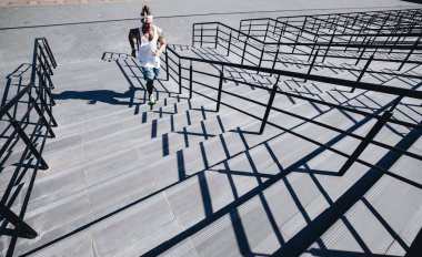 Beyaz t-shirt, siyah tayt ve mavi şort giymiş kafasında kafa bandı ile atletik adam güneşli bir günde merdivenlerden yukarı koşuyor