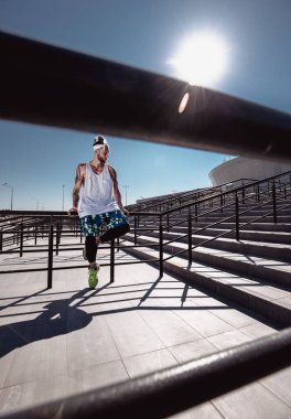 Yakışıklı kaslı delikanlı modern spor giyim oturma parlak güneşli bir günde, stadyum yanındaki basamaklarda eğitimden sonra