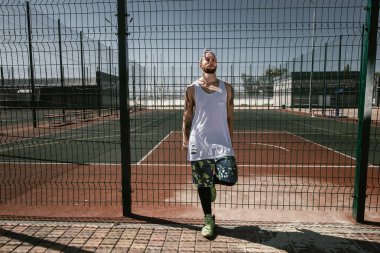 Atletik beyaz kafa bandı adamla beyaz t-shirt, siyah tayt giymiş ve güneşli bir günde Bahçesi çit yaslanmış duran mavi şort