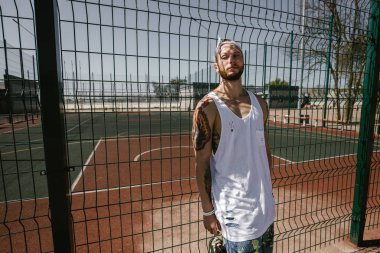 Beyaz kafa bandı ile genç adam beyaz t-shirt, siyah tayt giymiş ve güneşli bir günde Bahçesi çit yaslanmış duran mavi şort