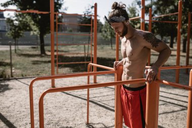 Atletik çıplak gövde ile kafa bandı erkekte siyah tayt giymiş ve kırmızı şort dışında spor zeminde asimetrik paralel yanındaki güneşli bir günde germe yapıyor