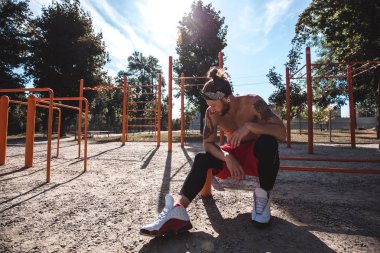 Kafa bandı çıplak gövde ile genç atletik adam siyah tayt giymiş ve kırmızı şort oturur spor ekipmanları spor zemin kum ile güneşli bir günde yakınındaki