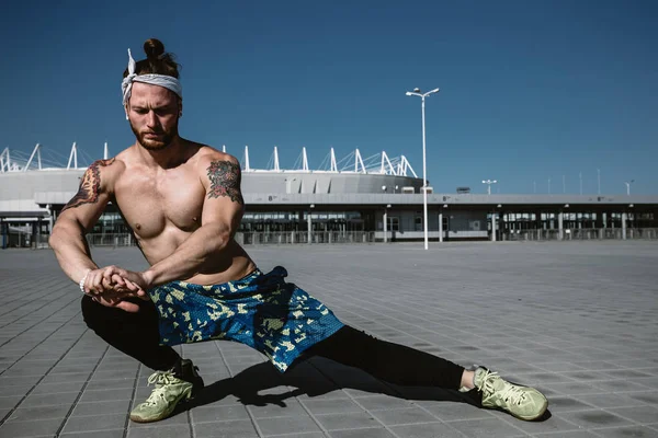 Jovem atlético com um tronco nu com a cabeça vestida com as pernas pretas e calções azuis está fazendo alongamento em lajes de pavimentação no fundo do estádio em um dia ensolarado quente — Fotografia de Stock