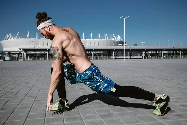 Jovem atlético com um tronco nu com a cabeça vestida com as pernas pretas e calções azuis está fazendo alongamento em lajes de pavimentação no fundo do estádio em um dia ensolarado quente — Fotografia de Stock