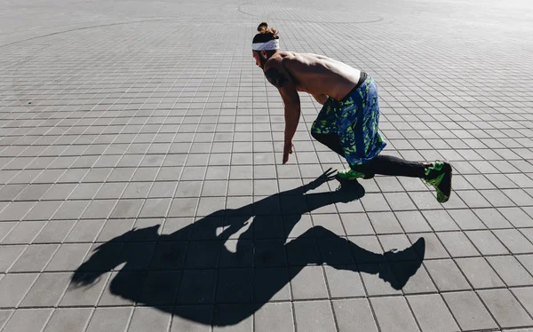 Jeune homme athlétique avec un torse nu avec des tatouages et un bandeau sur la tête habillé des leggings noirs et des shorts bleus commençant à courir sur les dalles de pavage sur la place par une chaude journée ensoleillée — Photo