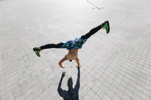 Homme athlétique avec un torse nu avec des tatouages habillés dans les leggings noirs et des shorts bleus faisant le tour debout sur une main sur les dalles de pavage dans la rue — Photo