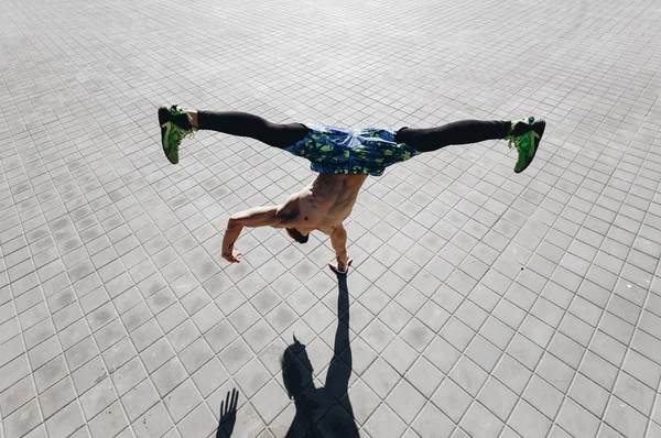 Homme athlétique avec un torse nu avec des tatouages habillés dans les leggings noirs et des shorts bleus faisant le tour debout sur une main sur les dalles de pavage dans la rue — Photo
