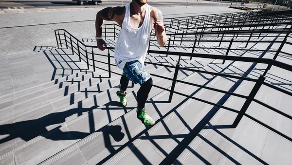 Atletische man met beugel op zijn hoofd gekleed in het wit t-shirt, zwarte legging en blauwe shorts loopt de trap buiten op een zonnige dag — Stockfoto