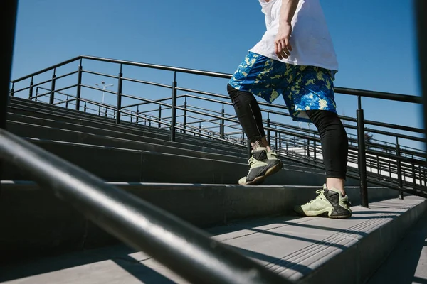 Athletic man with headband on his head dressed in the white t-shirt, black leggings and blue shorts is going up the stairs  with black railings outside on a sunny day