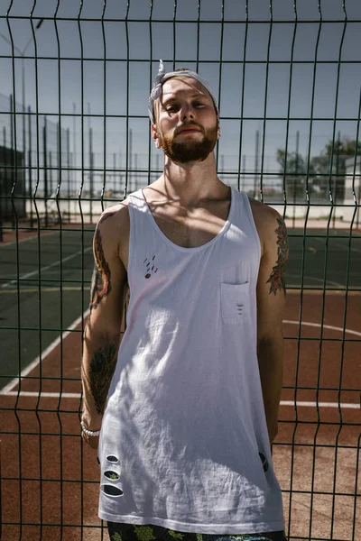 Young man with white headband dressed in the white t-shirt, black leggings and blue shorts stands leaning on the playground fence outside on a sunny day