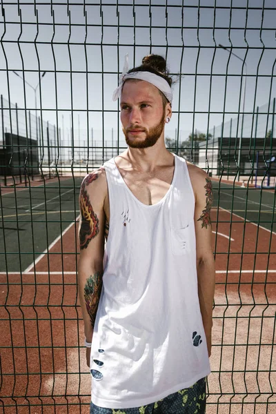 Young man with white headband  on his head and tattoos on his arms dressed in the white t-shirt, black leggings and blue shorts stands leaning on the playground fence outside on a sunny day