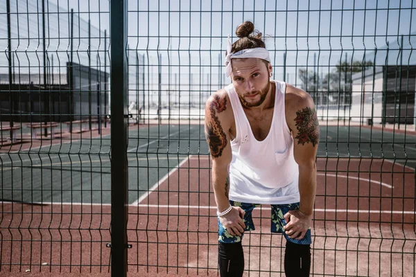 Jovem com cabeça branca na cabeça e tatuagens nos braços vestido com a camiseta branca, pernas pretas e shorts azuis fica ao lado da cerca do parque infantil do lado de fora em um dia ensolarado — Fotografia de Stock