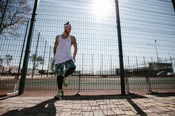 Jovem com cabeça branca na cabeça e tatuagens nos braços vestido com a camiseta branca, pernas pretas e shorts azuis se apoiam na cerca do parque infantil do lado de fora em um dia ensolarado — Fotografia de Stock