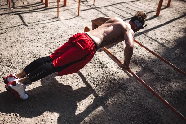 Jeune homme athlétique au bandeau avec un torse nu avec des tatouages vêtus de leggings noirs et des shorts rouges faisant des pompes sur les barres inégales à l'extérieur par une journée ensoleillée — Photo