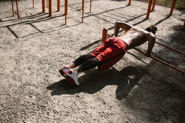 Jeune homme athlétique au bandeau avec un torse nu avec des tatouages vêtus de leggings noirs et des shorts rouges faisant des pompes sur les barres inégales à l'extérieur par une journée ensoleillée — Photo