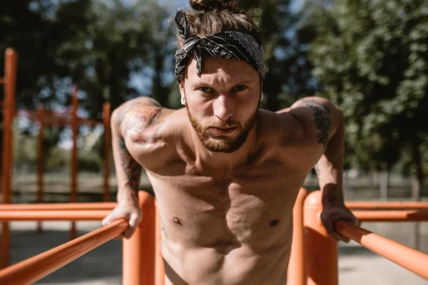 Jovem atlético na cabeça com um tronco nu com tatuagens fazendo flexões nos bares irregulares do lado de fora em um dia ensolarado — Fotografia de Stock