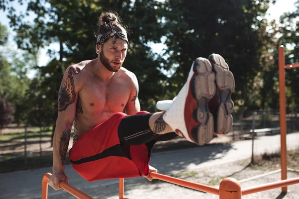 Young man in headband with naked torso dressed in black leggings and red shorts doing press exercises on the horizontal bar on the sports ground outside on a sunny day