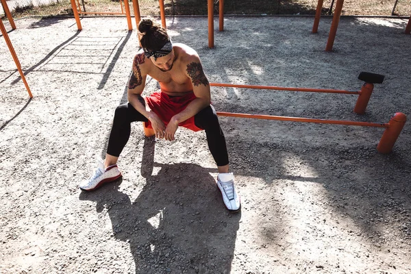 Young athletic man in headband with naked torso dressed in black leggings and red shorts sits near the sport equipment on the sports ground with sand on a sunny day