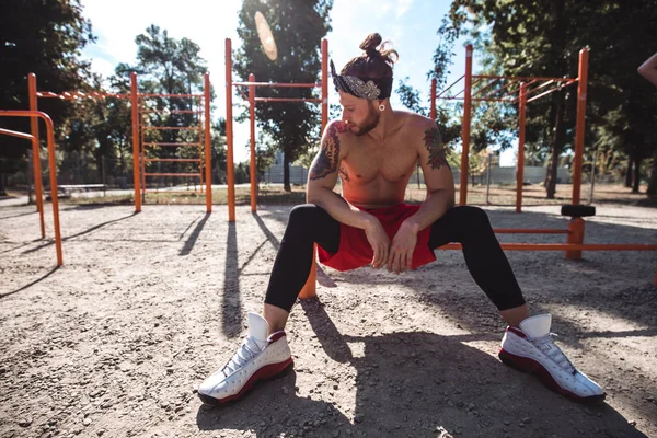 Young athletic man in headband with naked torso dressed in black leggings and red shorts sits near the sport equipment on the sports ground with sand on a sunny day