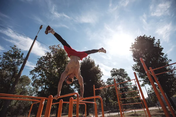 Homme athlétique en bandeau avec torse nu vêtu de leggings noirs et short rouge faisant le tour debout sur les mains sur la barre horizontale sur le terrain de sport à l'extérieur — Photo