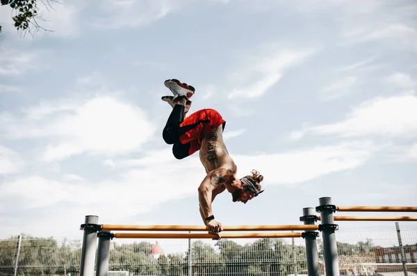 Jeune homme athlétique au bandeau avec torse nu vêtu de leggings noirs et de shorts rouges faisant un handstand sur des barres inégales sur le terrain de sport dehors par une journée ensoleillée — Photo