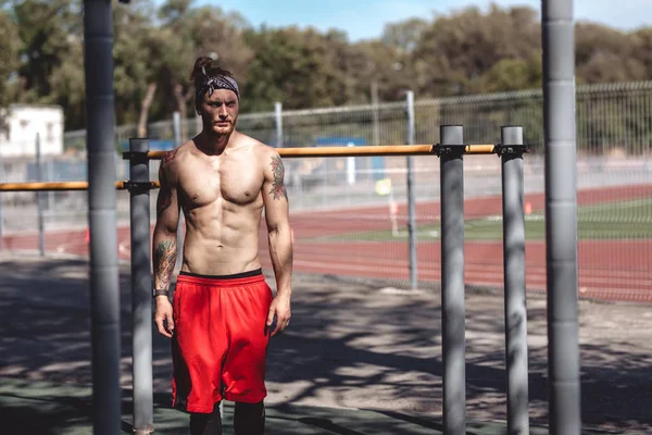 Athletic guy in headband with naked torso dressed in black leggings and red shorts stands on the sports ground outside next to the sports equipment